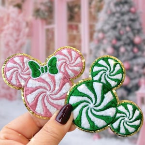 May include: Two embroidered patches in the shape of Mickey Mouse heads. One is pink and white with a green bow, the other is green and white. Both have gold glitter edging and a candy cane swirl design.