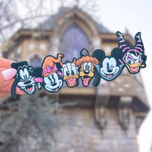 May include: A colorful patch featuring cartoon characters, including Mickey Mouse, Minnie Mouse, Donald Duck, and Goofy, with Halloween-themed hats. The patch is held up against a blurred background of a stone building.