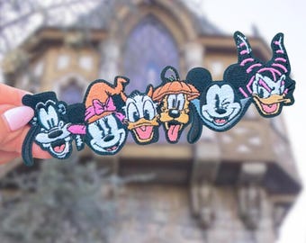 May include: A colorful patch featuring cartoon characters, including Mickey Mouse, Minnie Mouse, Donald Duck, and Goofy, with Halloween-themed hats. The patch is held up against a blurred background of a stone building.