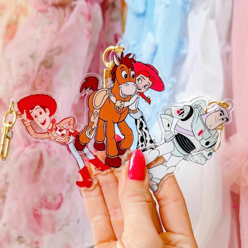 May include: Three acrylic keychains featuring characters from the Toy Story franchise. Woody in a cowboy outfit, Jessie hugging a horse, and Buzz Lightyear in a spacesuit. Each keychain has a gold-colored metal clasp.
