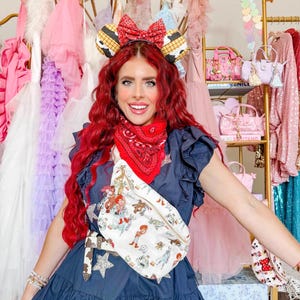 May include: A navy blue dress with star embellishments and a white fanny pack featuring a cartoon design. The fanny pack has a brown belt. The person is wearing a red bandana, a headband with a bow, and mouse ears.