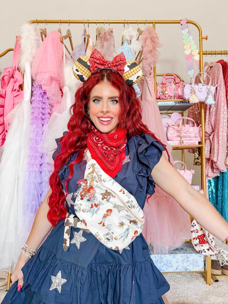 May include: A navy blue dress with star embellishments and a white fanny pack featuring a cartoon design. The fanny pack has a brown belt. The person is wearing a red bandana, a headband with a bow, and mouse ears.