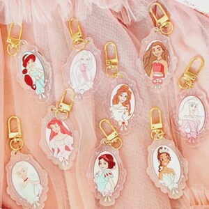 May include: A collection of clear acrylic keychains featuring various cartoon princess characters. Each keychain has a gold-colored clasp and a decorative oval frame. The princesses are depicted in their signature outfits and hairstyles, set against a pink background.