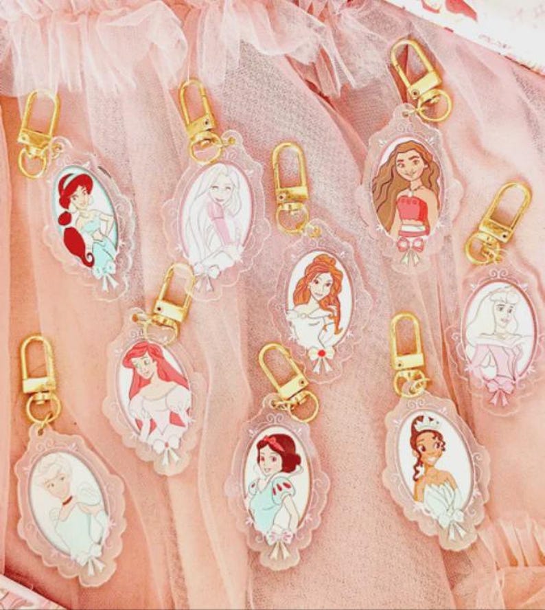 May include: A collection of clear acrylic keychains featuring various cartoon princess characters. Each keychain has a gold-colored clasp and a decorative oval frame. The princesses are depicted in their signature outfits and hairstyles, set against a pink background.