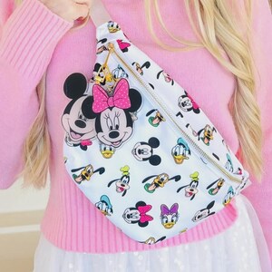 May include: White fanny pack featuring Mickey Mouse, Minnie Mouse, Donald Duck, Goofy, and Daisy Duck cartoon characters. The bag has a gold zipper and a pink bow charm. The strap is a light beige color.