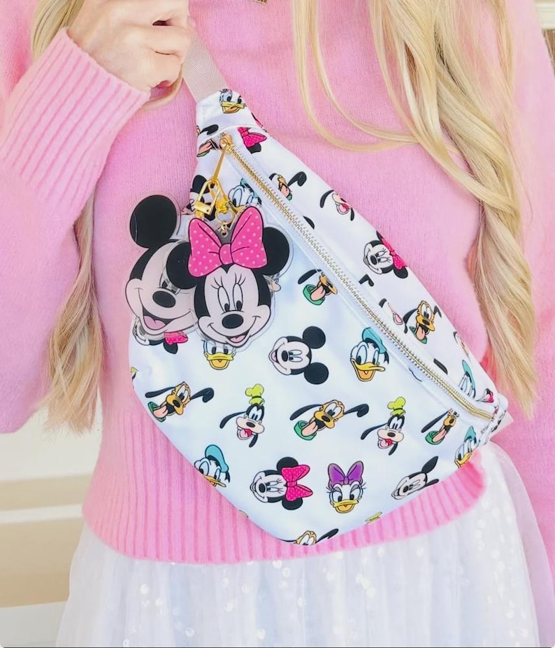 May include: White fanny pack featuring Mickey Mouse, Minnie Mouse, Donald Duck, Goofy, and Daisy Duck cartoon characters. The bag has a gold zipper and a pink bow charm. The strap is a light beige color.