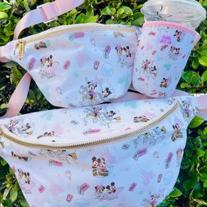 May include: White fanny packs and a drink holder with a pink strap and trim. The bags feature a repeating pattern of cartoon characters, castles, and other graphics. The drink holder has the text "PINK JOY CO".