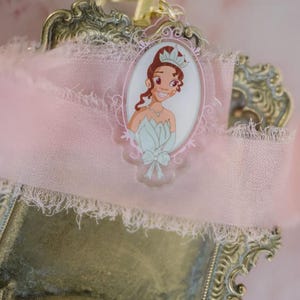 May include: A princess-themed keychain featuring a cartoon illustration of a princess in a light blue dress and crown. The keychain is set against a pink ribbon with a decorative, ornate, gold-colored metal frame.