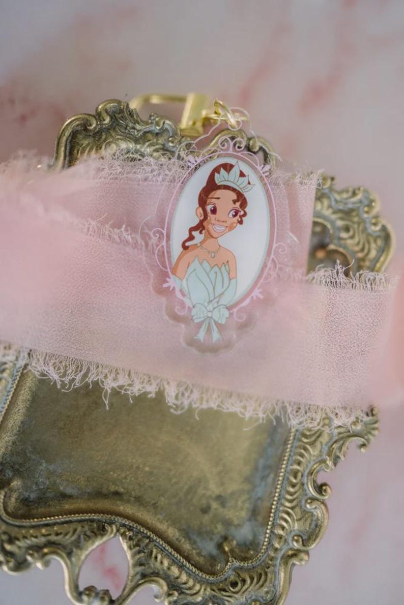 May include: A princess-themed keychain featuring a cartoon illustration of a princess in a light blue dress and crown. The keychain is set against a pink ribbon with a decorative, ornate, gold-colored metal frame.
