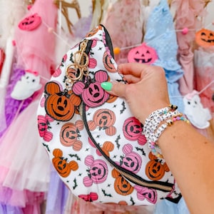 May include: A white belt bag featuring a repeating pattern of orange and pink Mickey Mouse-shaped jack-o'-lanterns and small black bats. The bag has a black zipper and two dangling charms shaped like orange and pink Mickey jack-o'-lanterns.