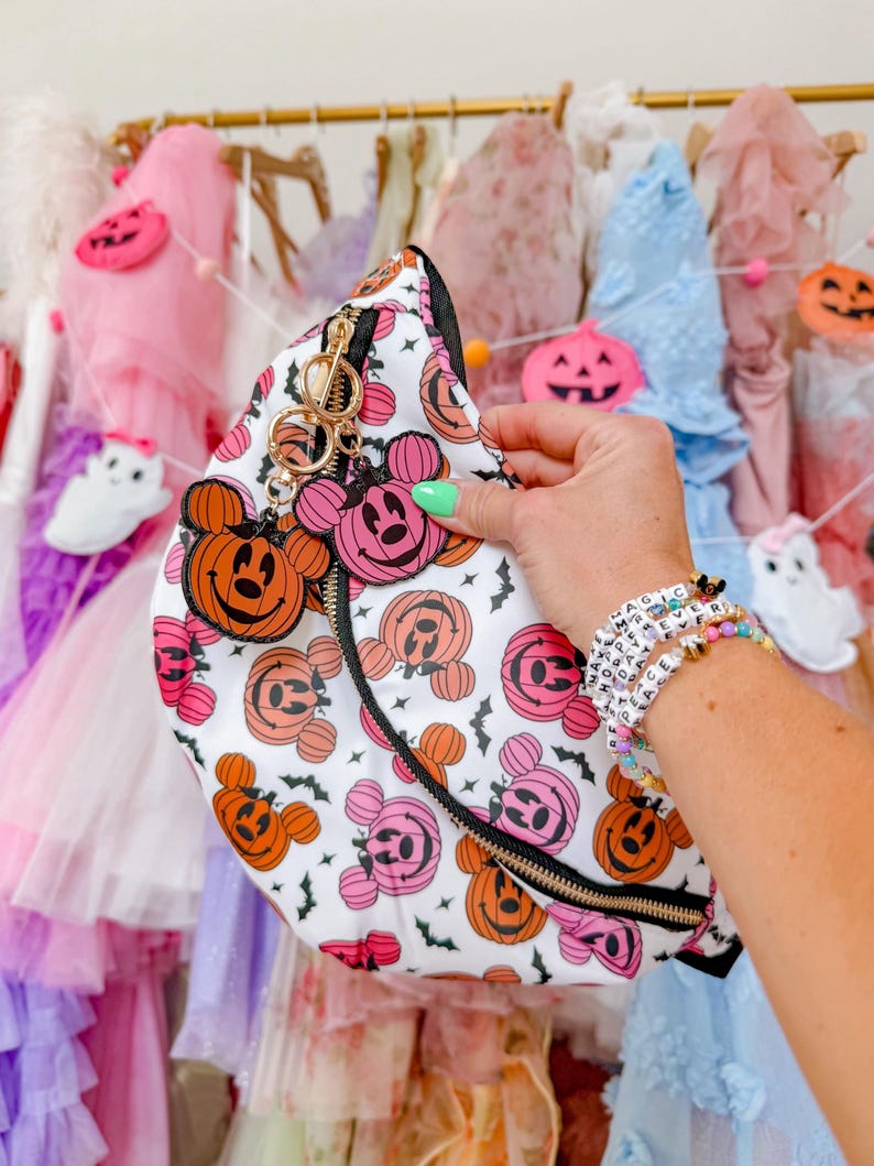 May include: A white belt bag featuring a repeating pattern of orange and pink Mickey Mouse-shaped jack-o'-lanterns and small black bats. The bag has a black zipper and two dangling charms shaped like orange and pink Mickey jack-o'-lanterns.