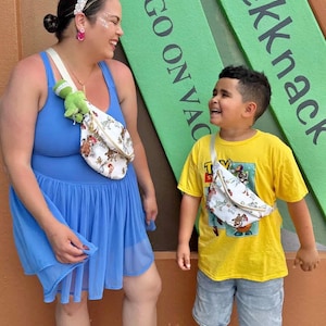 May include: Two people wearing fanny packs with a cartoon design. One person wears a blue dress and the other wears a yellow t-shirt and denim shorts. The background has green signs with white text that reads "SEE GO ON VAC" and "nickknack".
