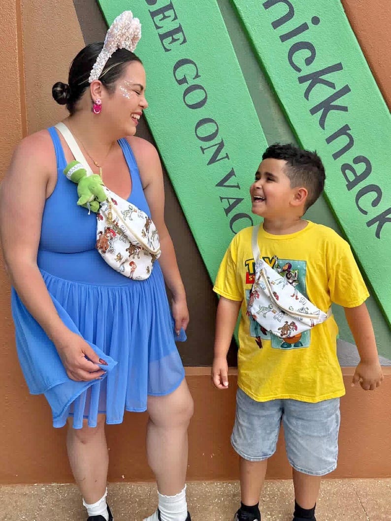 May include: Two people wearing fanny packs with a cartoon design. One person wears a blue dress and the other wears a yellow t-shirt and denim shorts. The background has green signs with white text that reads "SEE GO ON VAC" and "nickknack".
