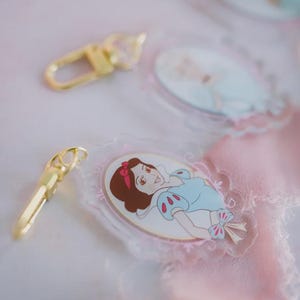 May include: Clear acrylic keychains featuring a cartoon image of Snow White in a blue dress with a red bow. The keychains have a gold-colored clasp. The background is a soft pink and white.