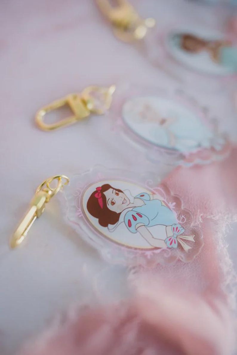 May include: Clear acrylic keychains featuring a cartoon image of Snow White in a blue dress with a red bow. The keychains have a gold-colored clasp. The background is a soft pink and white.