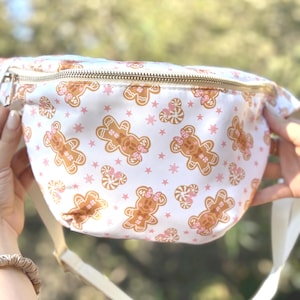 May include: A white fanny pack featuring a repeating pattern of brown gingerbread characters with pink bows, candy canes, and small pink stars. The bag has a gold-toned metal zipper closure across the top and an adjustable white strap.