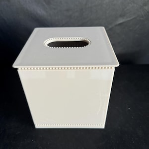 May include: A white square tissue box cover with a beaded trim and a rectangular opening for tissues.