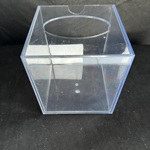 May include: A clear plastic square box with a lid and a circular opening on the top. The box is empty.
