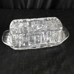 Op de afbeelding: Een glazen botervlootje van helder glas met een decoratief patroon. De vloot heeft een deksel en een rechthoekige vorm.