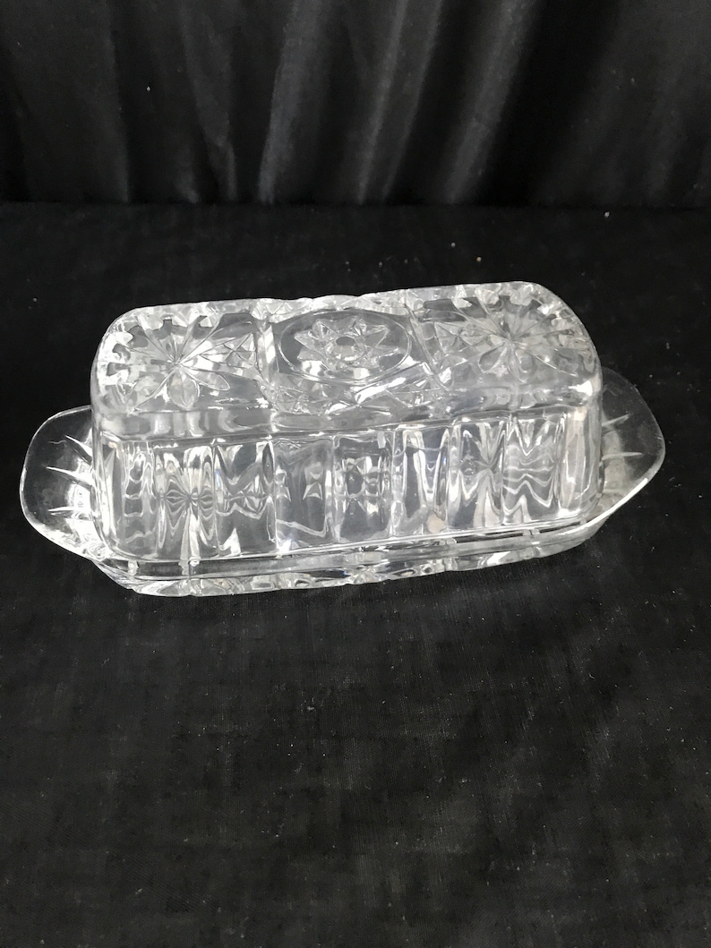 Op de afbeelding: Een glazen botervlootje van helder glas met een decoratief patroon. De vloot heeft een deksel en een rechthoekige vorm.