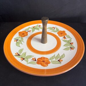 May include: A two-tiered serving tray with a central wooden handle. The tray features a white surface with orange floral and leaf designs, and an orange border. The handle is a dark brown color.
