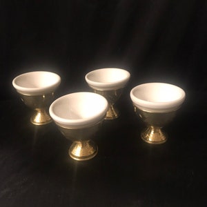 May include: Four small, decorative egg cups. Each cup has a white interior and a gold-colored exterior with a hammered texture. The cups are arranged on a dark surface, showcasing their elegant design.
