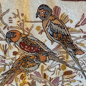 May include: A beige rug with an embroidered design of three birds with brown, gray, and orange feathers. The birds are perched on branches with brown and purple flowers.