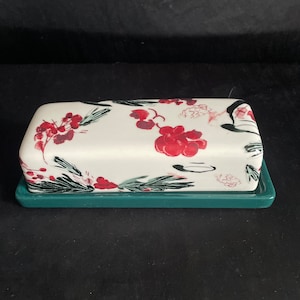 May include: A rectangular butter dish with a white lid featuring a floral design of red berries and green leaves. The base is a solid teal green. The dish is designed for serving butter.