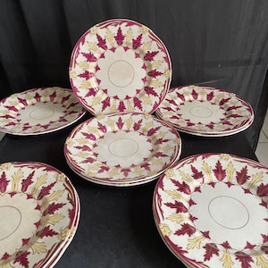 May include: A set of six antique plates with a cream-colored background. Each plate features a decorative border with burgundy and gold leaf patterns. The plates are arranged on a black surface, showcasing their intricate design and vintage charm.