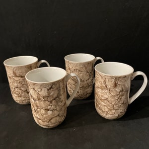 May include: Four ceramic mugs with a marbled pattern in shades of brown and cream. Each mug has a white interior and a curved handle. The mugs are arranged on a dark surface.
