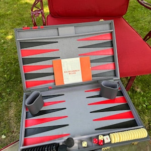 May include: Open backgammon set with gray interior, black, and red points. Includes dice, checkers, and a booklet titled "Backgammon Rules". The set is displayed on a glass table with a red chair in the background.