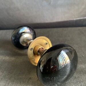 May include: Close-up of two antique black glass door knobs with brass hardware. The knobs have a glossy finish and reflect the surrounding environment. The hardware shows signs of age and wear, adding to their vintage appeal.