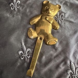 May include: A brass teddy bear wall hook. The hook is in the shape of a bear with a bow tie, and a long hook extending from the bottom. The hook is made of brass and is designed to be mounted on a wall. The background is a dark gray fabric.