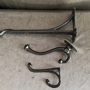 May include: Three black cast iron hooks and a shelf bracket. The shelf bracket is long and has a curved arm. Two of the hooks have a curved design, and the third is smaller with a right-angle design.