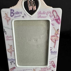 May include: A white ceramic picture frame with a pink and purple ballet dancer theme. The frame has a heart-shaped cutout at the top with a pair of pink ballet shoes hanging from it. The frame is decorated with ballet dancers, music notes, and the words "Ballet Dancer".