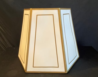 May include: A white lampshade with a geometric design. The lampshade has a trapezoidal shape with a gold border and trim. The white panels are rectangular and the gold trim outlines the panels. The lampshade is sitting on a black surface.