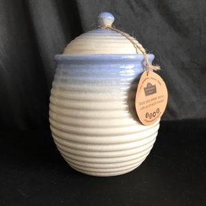 May include: A ceramic jar with a lid, featuring a light blue and white color scheme. The jar has a ribbed design and a small, round knob on top. A tag is attached with twine.