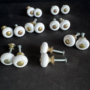 May include: A collection of 16 white ceramic cabinet knobs with gold accents. The knobs have a round shape and a simple design.