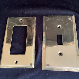 May include: Two shiny, brass-colored electrical switch plates. One plate has a rectangular opening, and the other has a single switch opening. Both plates have screw holes for mounting. The plates are on a dark blue surface.