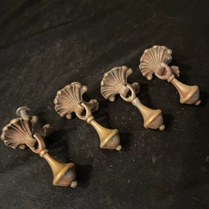 May include: Four antique bronze-toned drawer pulls. Each pull features a fan-shaped top, a decorative stem, and a small, round base. The pulls are arranged on a dark, textured surface, showcasing their vintage design and potential for furniture restoration.