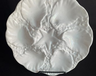 May include: White ceramic oyster serving platter with six compartments and a central star-shaped design. The platter has a scalloped edge and a textured, embossed surface. A decorative piece for serving seafood.