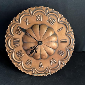 May include: A copper wall clock with Roman numeral markings and a decorative, embossed design around the edge. The clock face has a raised, textured center.