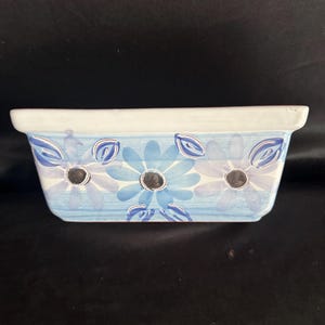 May include: A rectangular ceramic planter with a blue and white floral design. The planter has three drainage holes in the bottom.