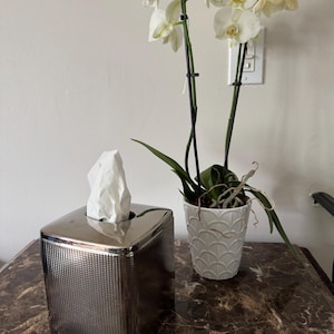 May include: A silver tissue box with a textured pattern sits on a marble-patterned surface. A white orchid in a white scalloped pot is next to the tissue box. The orchid has white flowers and green leaves.