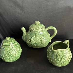 May include: A green ceramic tea set with a teapot, creamer, and sugar bowl. Each piece features a raised design of rabbits and flowers.