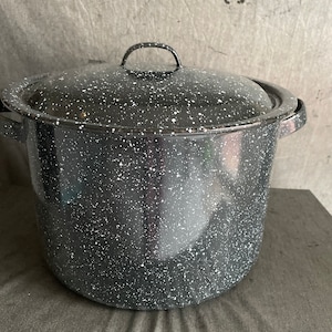 May include: A large, speckled black and white enamel pot with a matching lid. The pot has two side handles and a loop handle on the lid. The pot is sitting on a gray surface.
