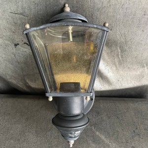 May include: A black outdoor wall-mounted lantern with a clear glass enclosure. The lantern has a decorative top and bottom, with a small finial on top. The glass is slightly tinted, and the fixture appears to be made of metal.