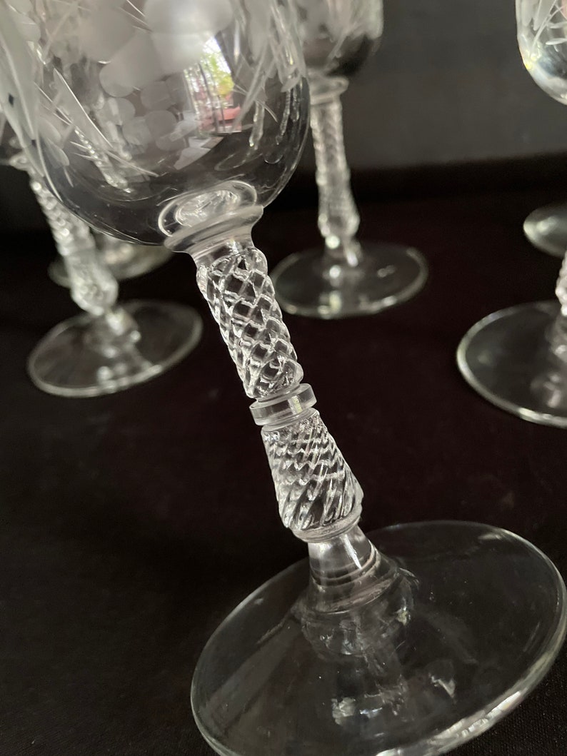May include: Clear glass wine glasses with etched floral designs. The stems have a diamond-cut pattern. The glasses are on a dark surface. The image shows a close-up of the glasses.