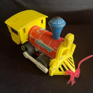 May include: A vintage toy train with a yellow cab and front, a red body with the text "WOOT-WOOT", and a blue smokestack. The train has black wheels and a white connecting rod. A pink string is attached to the front.