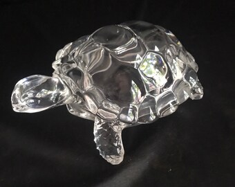 May include: A clear, faceted glass turtle ornament. The turtle has a detailed shell, head, and legs. The glass reflects light, creating a sparkling effect. The ornament is a decorative item.